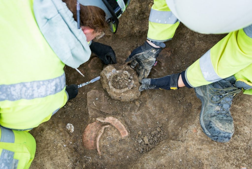 Two archaeologist carefully lifting a whole pottery vessel from a small, shallow, excavated pit. In the pit is another vessel, partially excavated